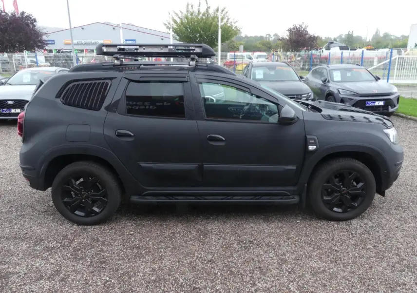 Profil côté gauche du Dacia Duster 2023 noir mat avec galerie de toit, marchepied et jantes noires.