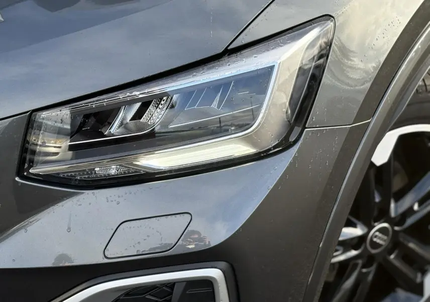 Gros plan sur le phare avant droit et la jante Audi Sport 5 branches titane mat du Q2 gris Daytona.