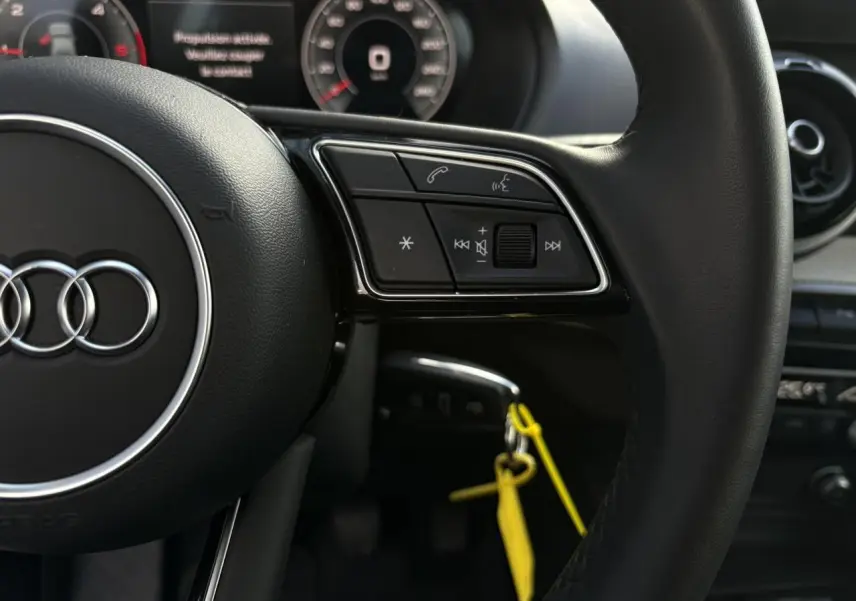 Gros plan sur le volant multifonction en cuir noir de l'Audi Q2 30 TDI S-Line, avec commandes audio et tableau de bord partiel.