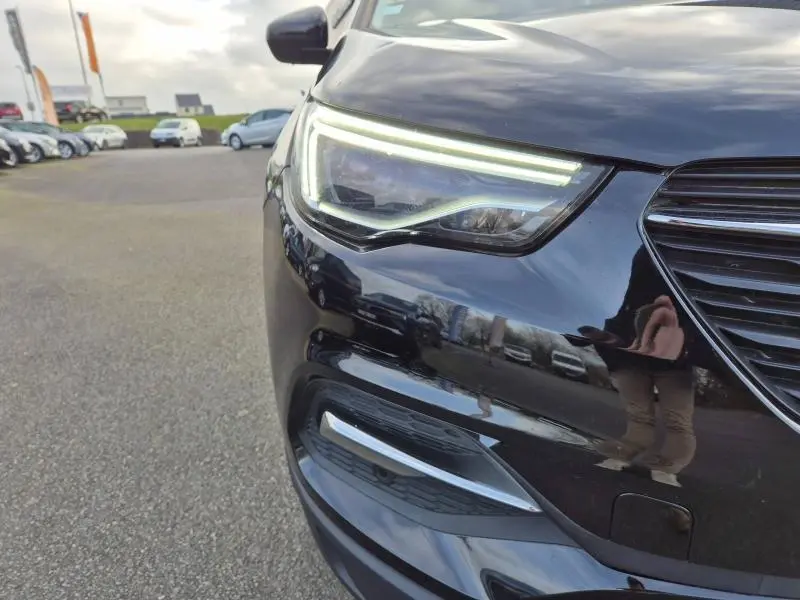 Gros plan sur le phare avant gauche allumé d’un Opel Grandland X noir diamant, avec calandre et rétroviseur visibles.