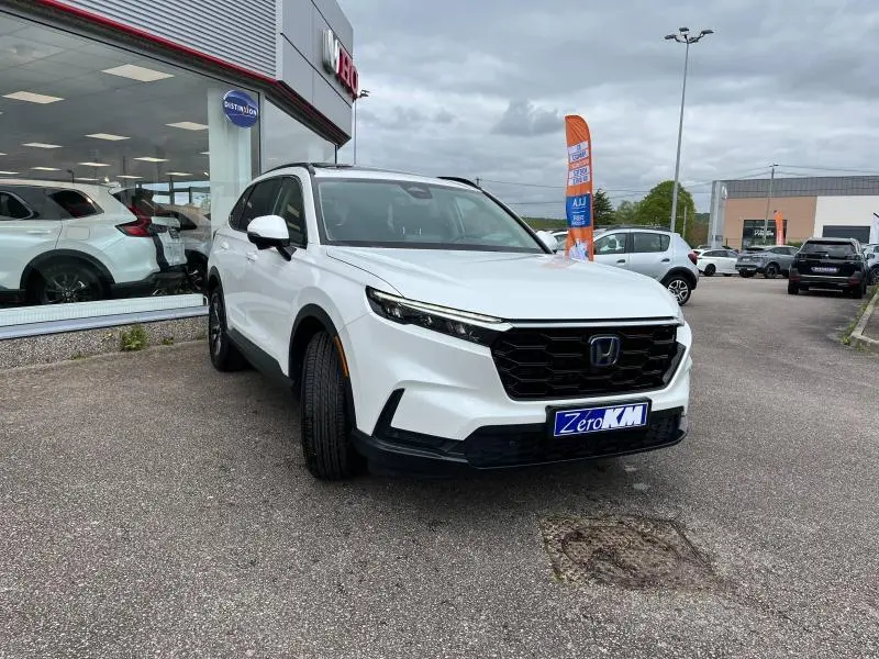 Vue 3/4 avant d'un Honda CR-V 2024 blanc platine nacré, avec calandre noire et jantes alliage grises, stationné devant un showroom.