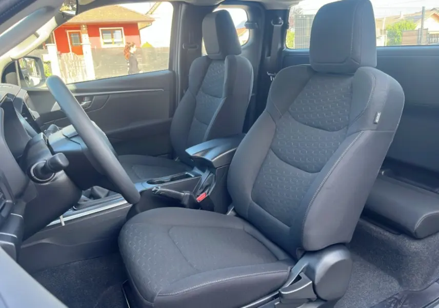 Vue intérieure côté gauche du poste de conduite de l'Isuzu D-MAX 2026 avec sièges tissu gris foncé et accoudoir central.