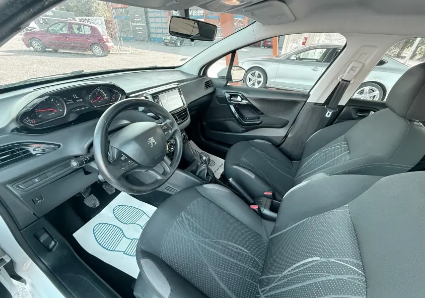 Vue intérieure côté conducteur d'une Peugeot 208 blanche de 2013, montrant le volant, tableau de bord et sièges noirs.