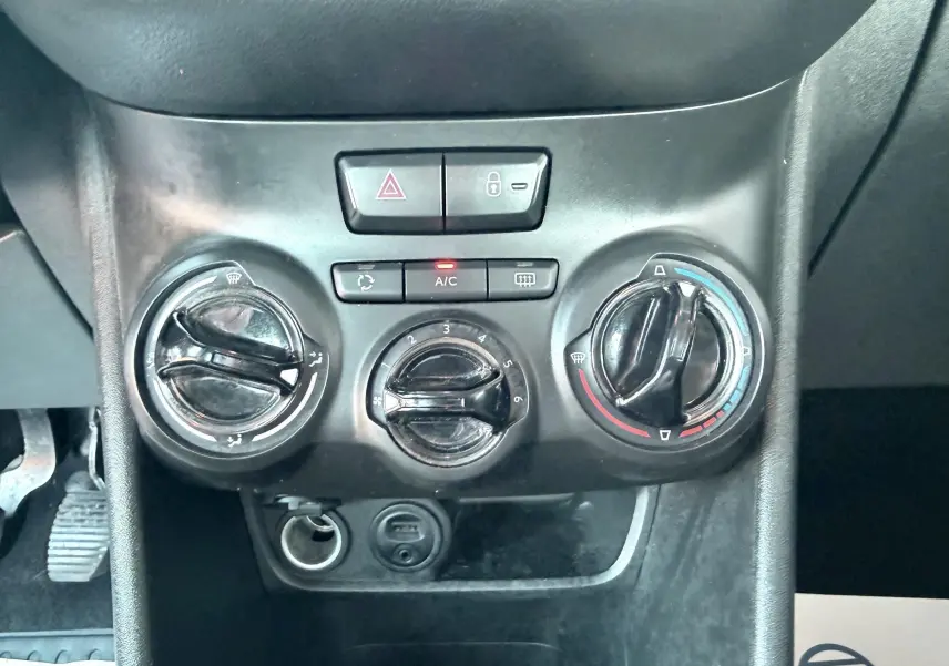 Gros plan sur la console centrale noire de la Peugeot 208 blanche de 2013, montrant les commandes de climatisation et boutons d’alerte.