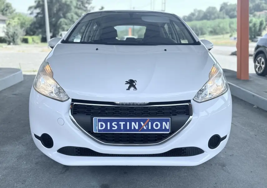 Vue frontale d'une Peugeot 208 blanche de 2013 avec calandre noire et phares allumés, plaque Distinxion visible.