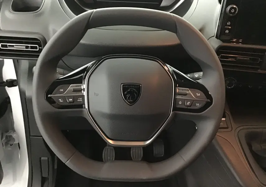 Vue rapprochée du volant noir du Peugeot Partner blanc, avec commandes intégrées et tableau de bord sombre en arrière-plan.