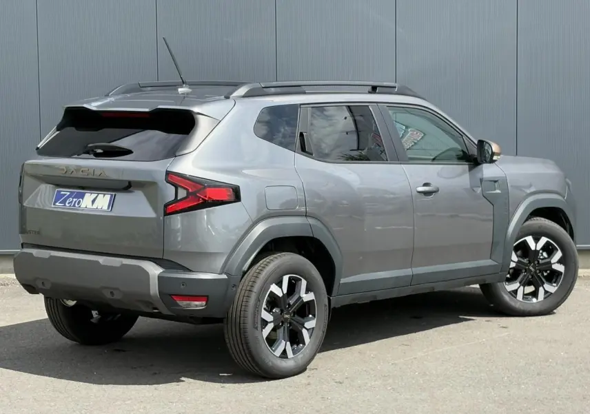Vue 3/4 arrière droite du Dacia Duster 2025 gris schiste avec jantes alliage et barres de toit noires.