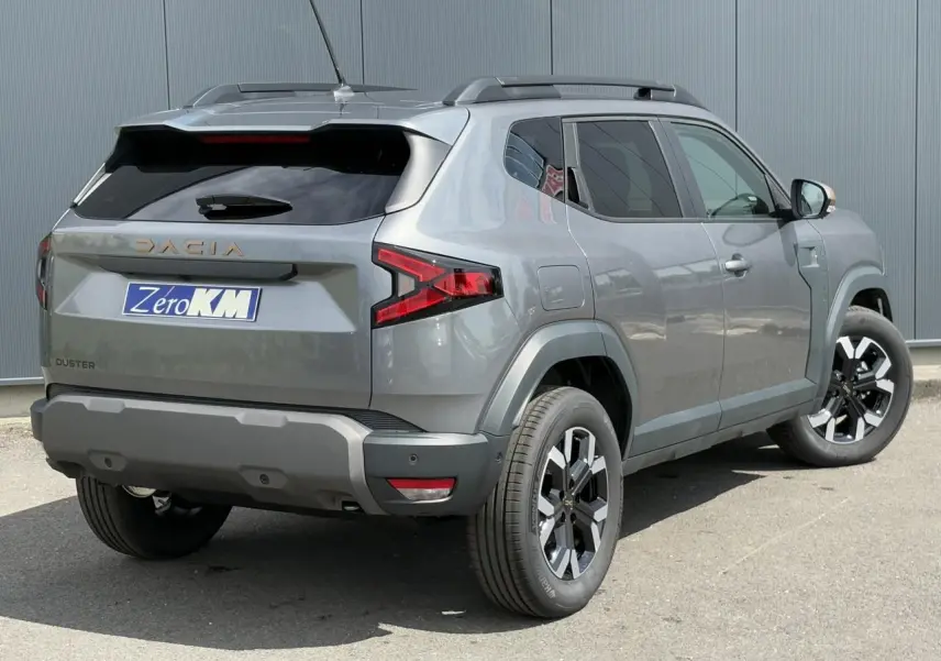 Vue 3/4 arrière droit du Dacia Duster gris schiste avec jantes alliage et barres de toit noires.