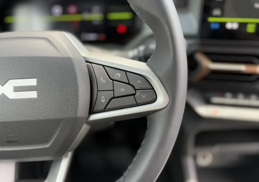 Gros plan sur le volant gris du Dacia Duster 2025 avec commandes vocales et écran digital flou en arrière-plan.