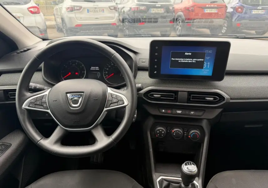Vue intérieure du tableau de bord et volant noir d'une Dacia Sandero blanche, avec écran tactile et boîte manuelle visible.