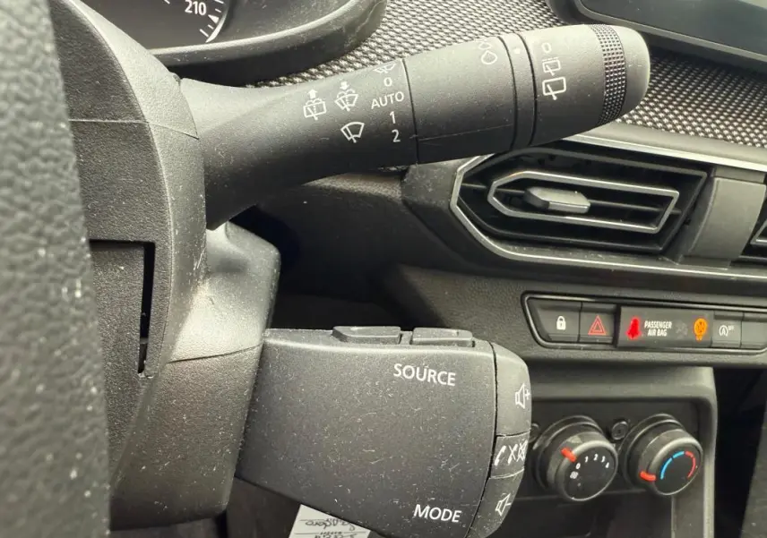 Gros plan sur la commande de source audio et les réglages de climatisation dans le tableau de bord noir d'une Dacia Sandero blanche.