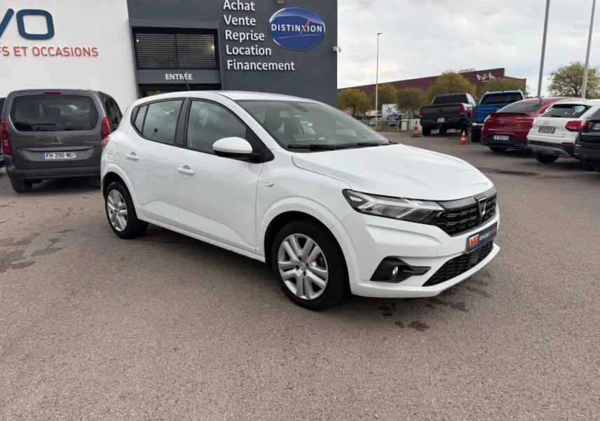 Dacia Sandero blanc vue 3/4 avant droit, garée devant un bâtiment commercial avec plusieurs autres voitures visibles.
