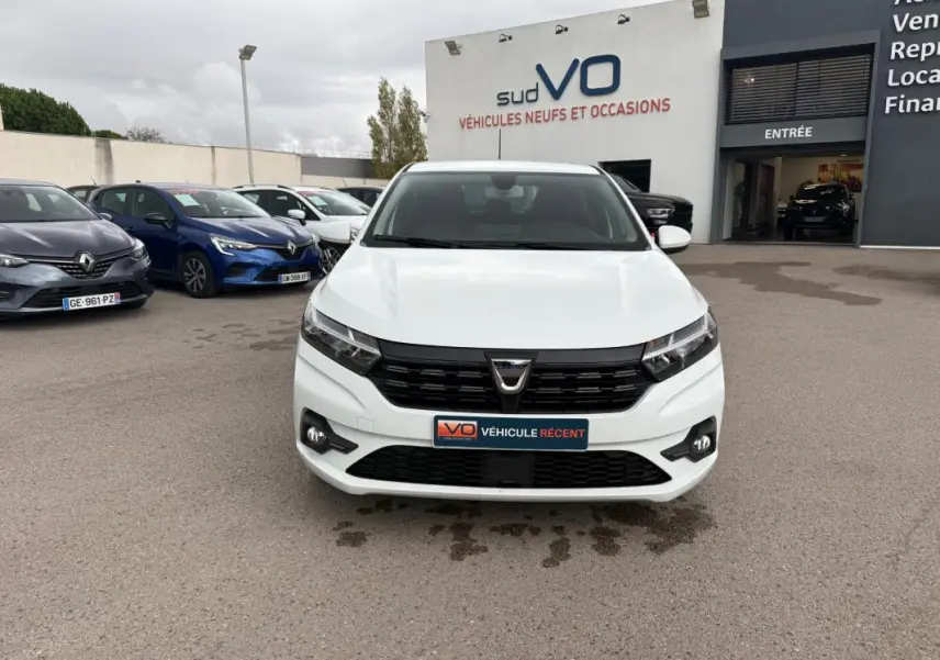Vue frontale d'une Dacia Sandero blanche 2021 avec calandre noire et feux antibrouillard sur un parking de concession.