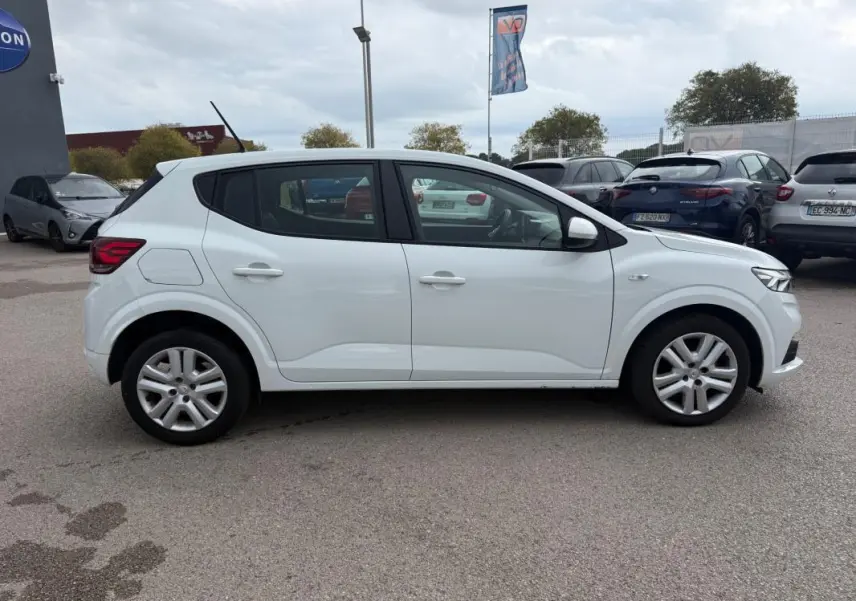 Profil droit d'une Dacia Sandero blanche 2021 avec vitres surteintées et jantes alliage en arrière-plan de parking.