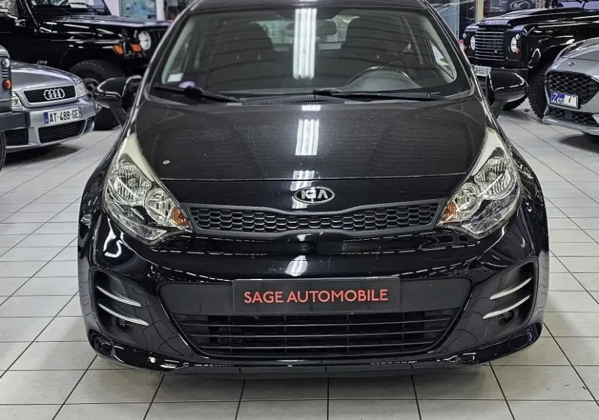 Vue frontale d'une KIA RIO III noire de 2015, exposée en intérieur avec plaque "SAGE AUTOMOBILE" visible.
