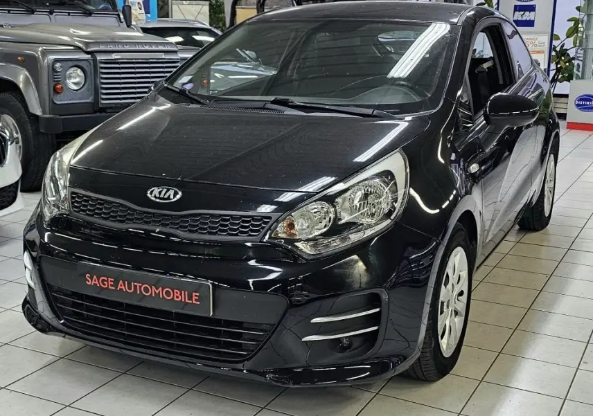 Vue 3/4 avant d'une KIA RIO III noire de 2015, compacte 3 portes avec phares larges et jantes blanches en showroom.