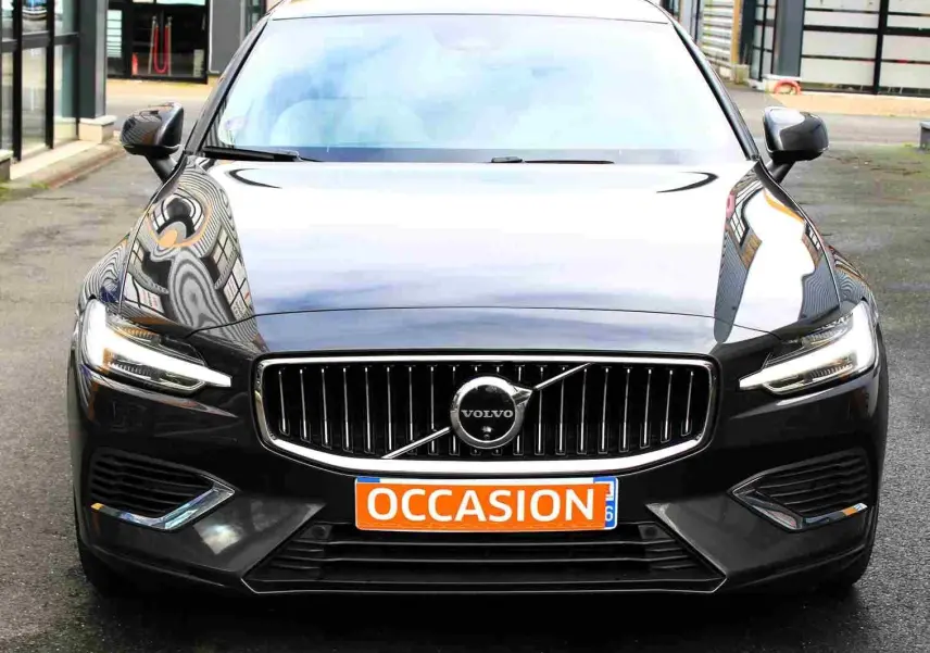 Vue frontale d'une Volvo S60 gris métal 2024 avec calandre chromée et phares LED allumés, plaque "OCCASION" visible.