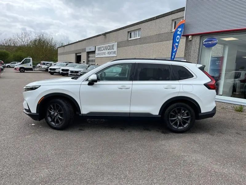 Vue de profil côté gauche du Honda CR-V 2024 blanc platine nacré avec jantes alliage noires, stationné devant un atelier mécanique.