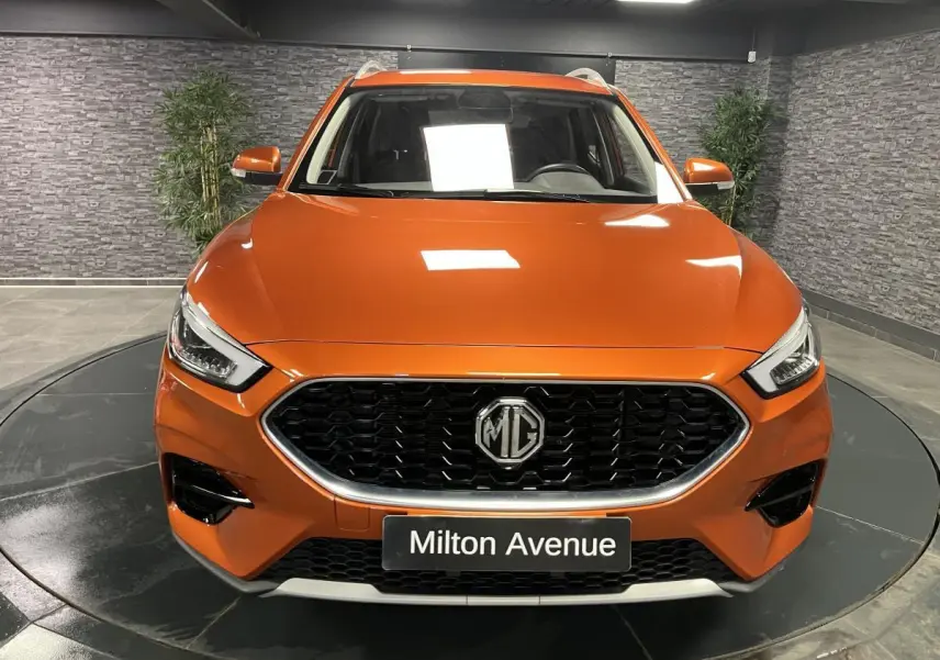 Vue frontale d'un MG ZS orange métallisé avec calandre noire et logo MG bien visible, en intérieur showroom.