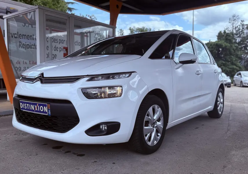 Citroën C4 Picasso blanc vue 3/4 avant droit, avec calandre noire et phares allumés sous un abri orange.