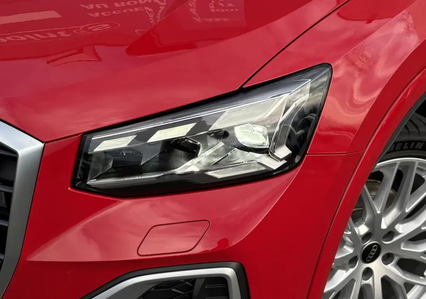 Gros plan sur le phare avant droit et la roue argentée d’un Audi Q2 rouge métallisé 2025 finition S-Line.