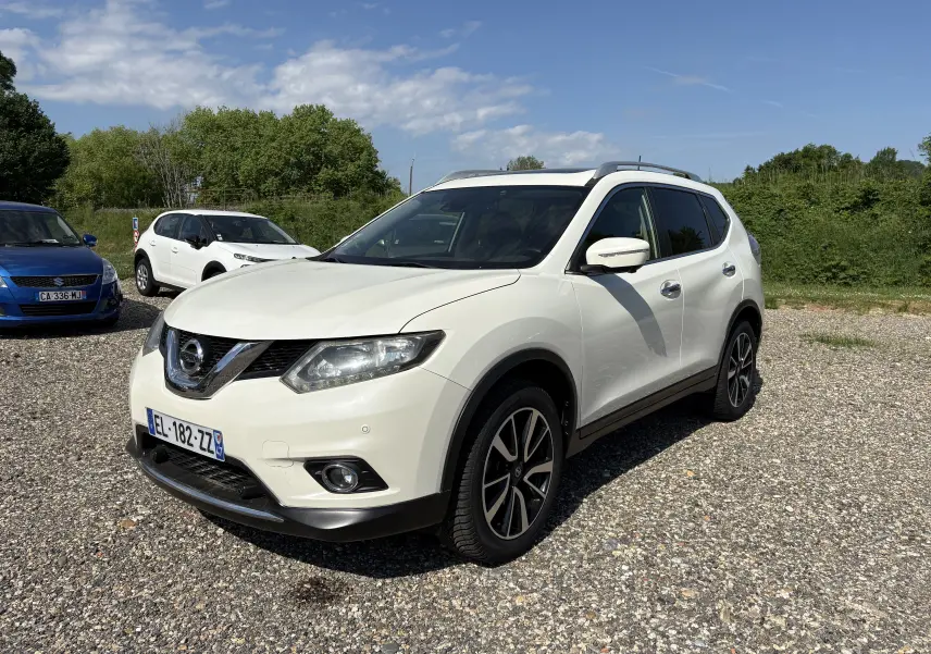 Nissan X-Trail 2016 blanc en 3/4 avant droit, avec jantes alliage et barres de toit visibles sur un sol gravillonné.