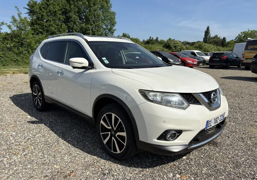 Nissan X-Trail 2016 blanc en 3/4 avant droit, avec jantes alliage et barres de toit visibles sur parking extérieur.