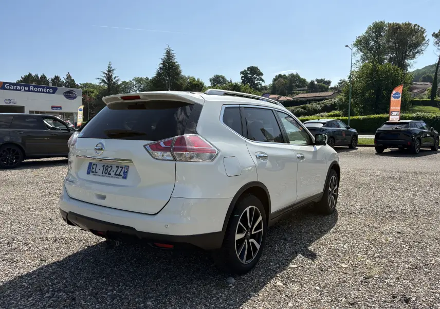Nissan X-Trail blanc 2016 en 3/4 arrière droit, avec jantes alliage et vitres teintées, stationné sur un parking extérieur.