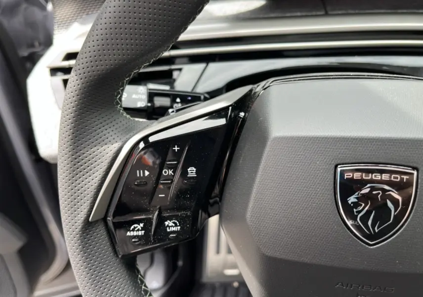 Gros plan sur le volant cuir perforé du Peugeot 5008 Hybrid GT avec commandes multifonctions et logo lion noir brillant.
