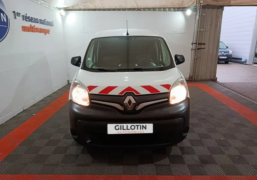 Vue avant d’un Renault Kangoo blanc 2020 avec bandes rouges diagonales sur le capot et phares allumés dans un garage.