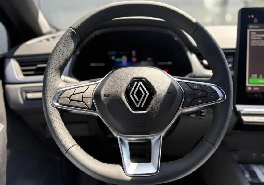 Volant cuir multifonctions noir avec logo Renault au centre, tableau de bord et écran tactile du Captur hybride 2025 visibles en arrière-plan.