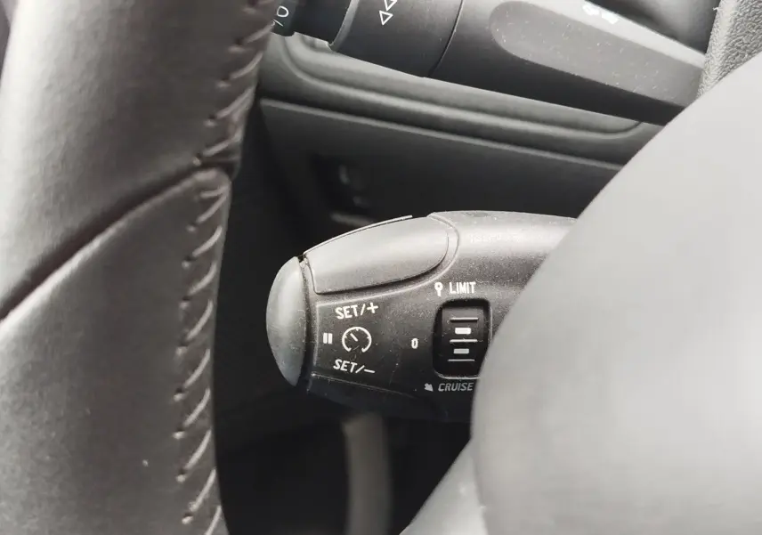 Gros plan sur le commodo de régulateur de vitesse à gauche du volant d’une Peugeot 208 noire, intérieur sombre.