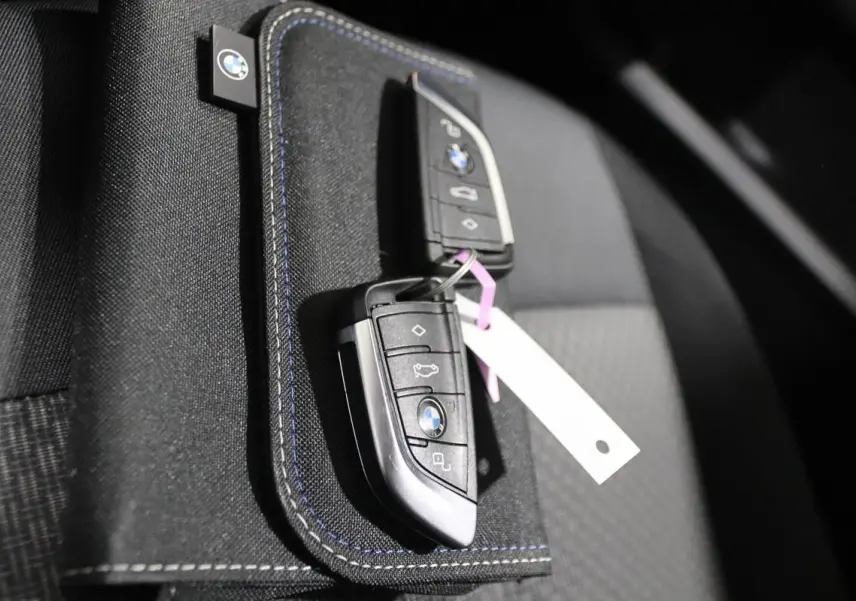 Gros plan sur deux clés BMW posées sur un porte-clés en tissu noir avec surpiqûres bleues, intérieur de siège gris.