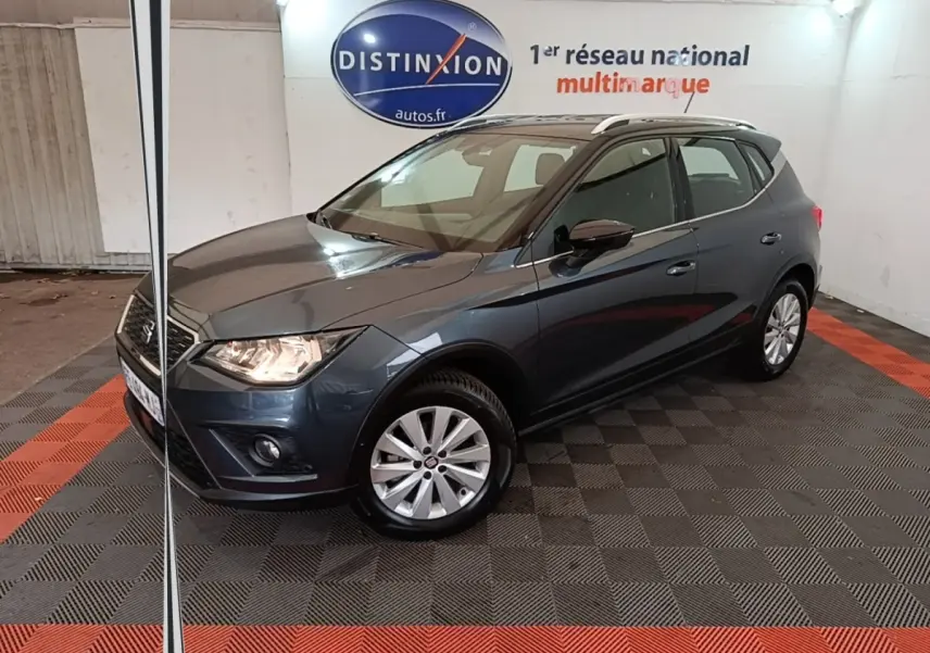 SEAT Arona gris clair en 3/4 avant droit, avec jantes alliage et feux allumés dans un showroom intérieur.