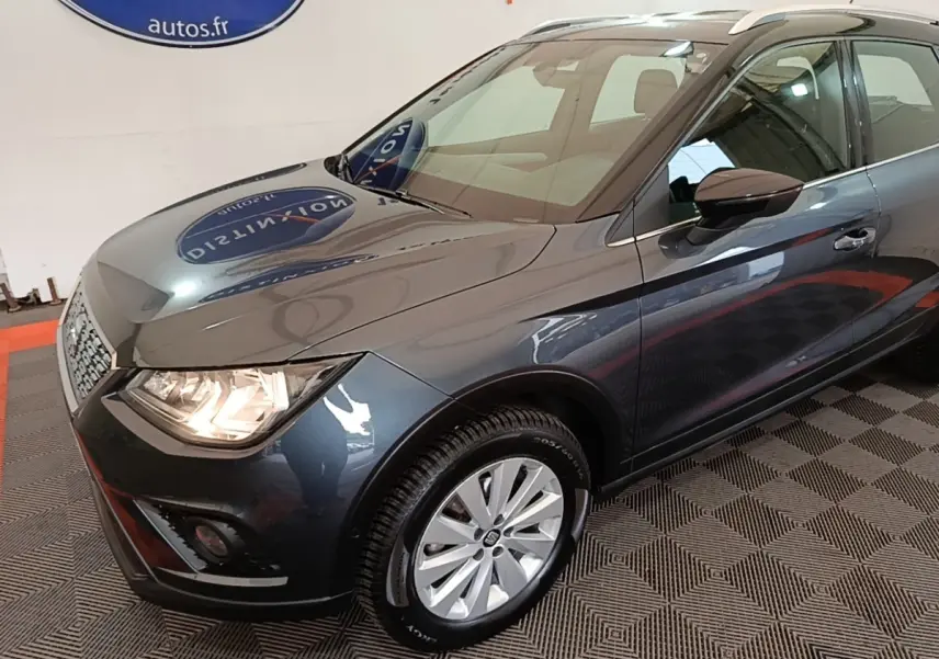 SEAT Arona gris clair vue 3/4 avant droit, phares allumés et jantes alliage visibles en intérieur showroom.