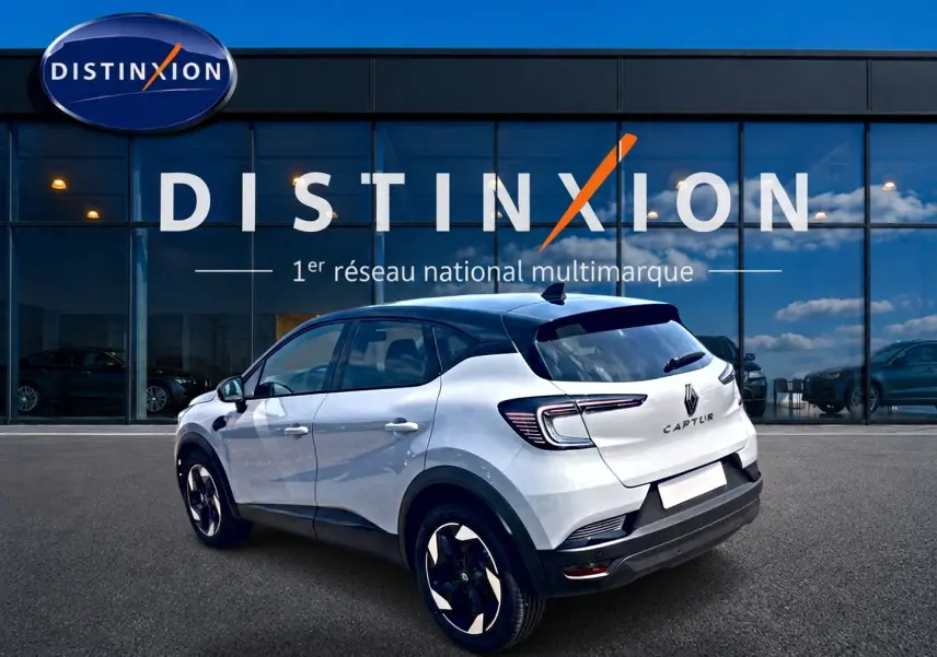 Vue 3/4 arrière droite du Renault Captur 2026 blanc Rafale avec toit noir et jantes alu noires devant un bâtiment vitré Distinxion.