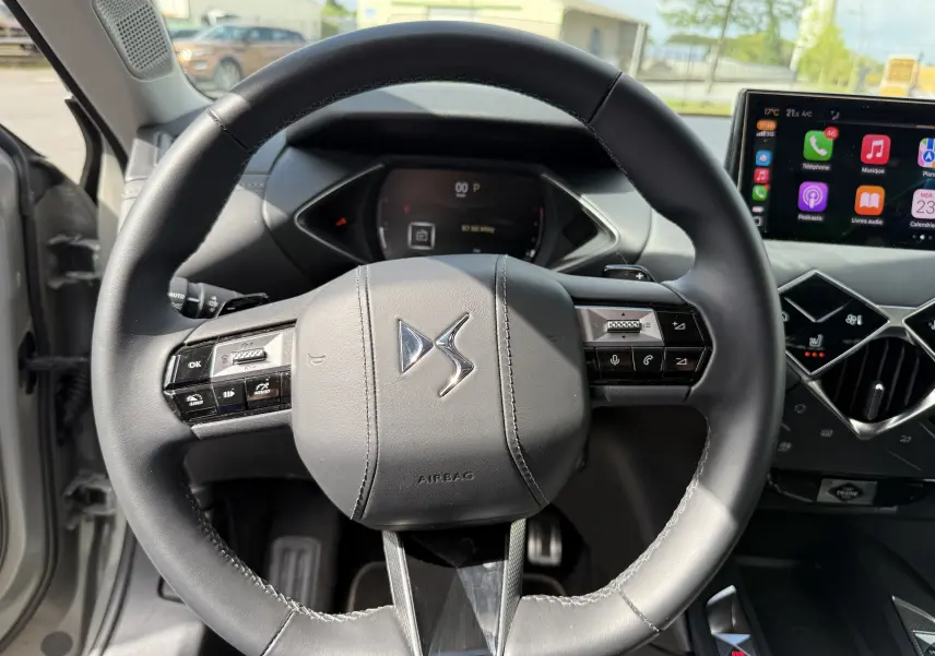 Vue rapprochée du volant cuir noir du DS3 2024 avec commandes intégrées et tableau de bord numérique en arrière-plan.