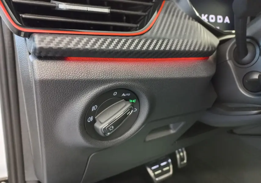 Gros plan sur le bouton de commande des phares avec finition carbone et éclairage d'ambiance rouge dans un Skoda Kamiq Blanc Lune.