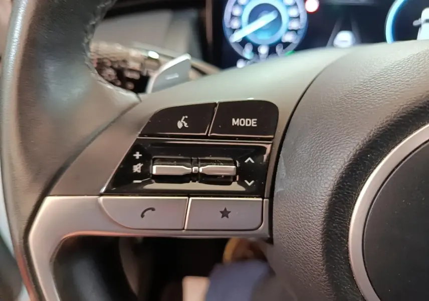 Gros plan sur les commandes gauche du volant cuir noir du Hyundai Tucson blanc hybride 2022 avec tableau de bord numérique.