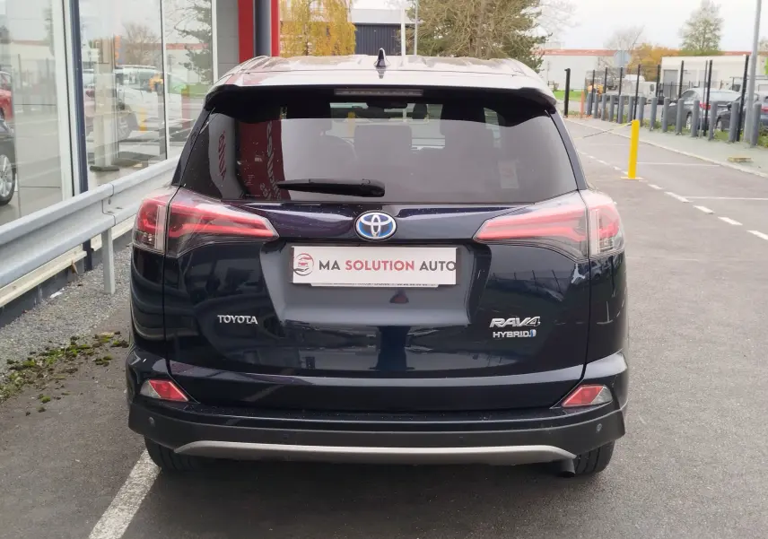 Vue arrière d'un Toyota RAV4 Hybride noir cobalt garé devant un bâtiment vitré, avec logo et feux LED visibles.