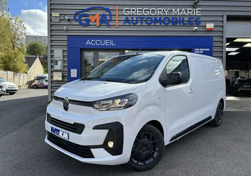 Vue 3/4 avant d’un Citroën Jumpy blanc stationné devant un garage avec jantes noires et phares allumés.