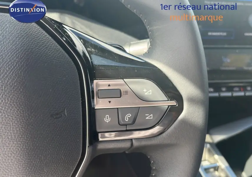 Gros plan sur les commandes gauche du volant cuir noir de la Peugeot 308 Hybrid 2025, avec tableau de bord en arrière-plan.