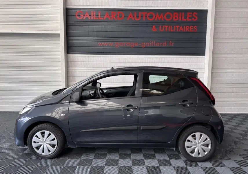 Profil côté gauche d'une Toyota Aygo 1.0 VVT-i x-black gris foncé, avec jantes standard et intérieur visible.