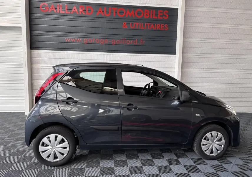 Profil côté gauche d'une Toyota Aygo 1.0 VVT-i x-black gris foncé, en intérieur sur sol damier, avec jantes argentées.