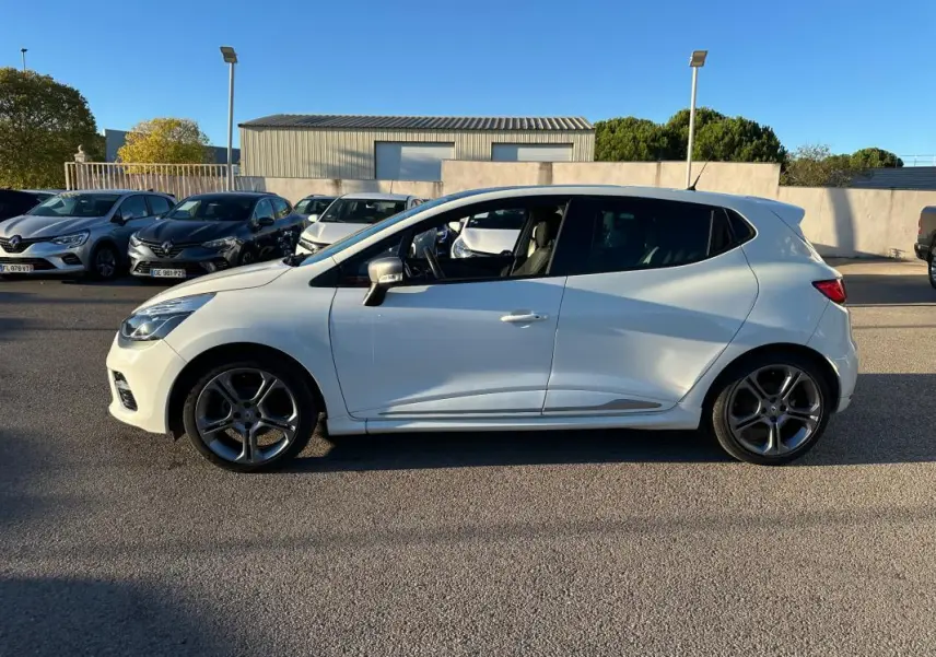 Profil côté gauche d'une Renault Clio IV blanche avec jantes alliage 17 pouces sur fond de parking ensoleillé.