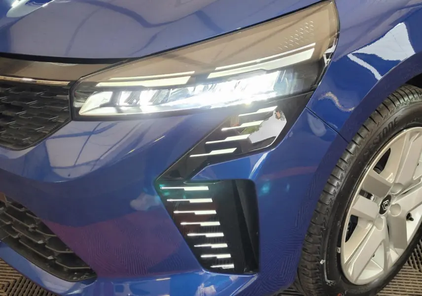 Gros plan sur le phare avant droit allumé d'une Renault Clio 2025 bleu avec jante argentée et détails LED distinctifs.