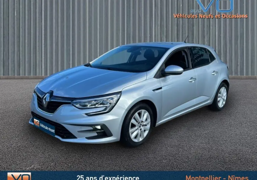 Renault Mégane IV Berline gris clair vue 3/4 avant droit sur sol bitumé devant un mur métallique