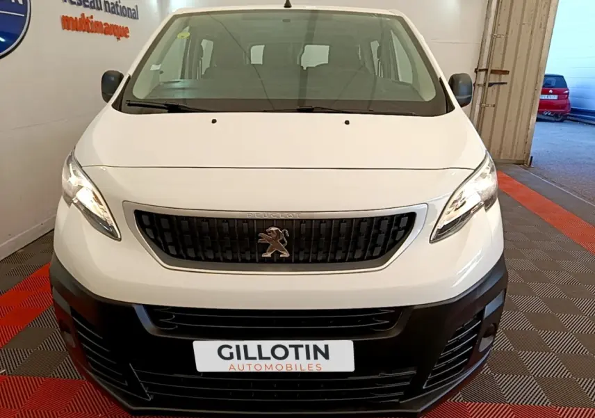 Vue avant d’un Peugeot Traveller blanc de 2018 avec calandre noire et logo lion visible, stationné en intérieur.