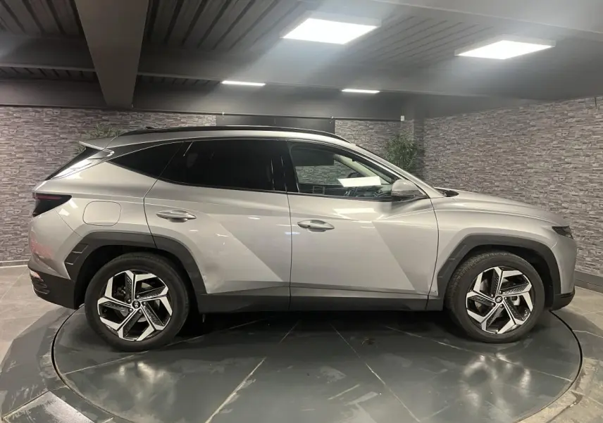 Profil droit du Hyundai Tucson 2023 gris métallisé avec jantes alliage et vitres surteintées en intérieur showroom.