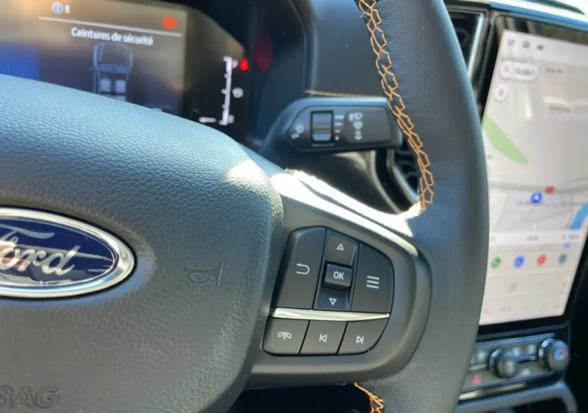 Gros plan sur le volant noir avec surpiqûres orange du Ford Ranger 2025, affichage numérique et écran tactile visible.